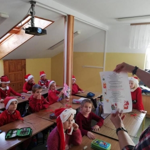 Nauczycielka odczytuje list od Mikołaja, w ławkach siedzą uczniowie w mikołajkowych czapkach