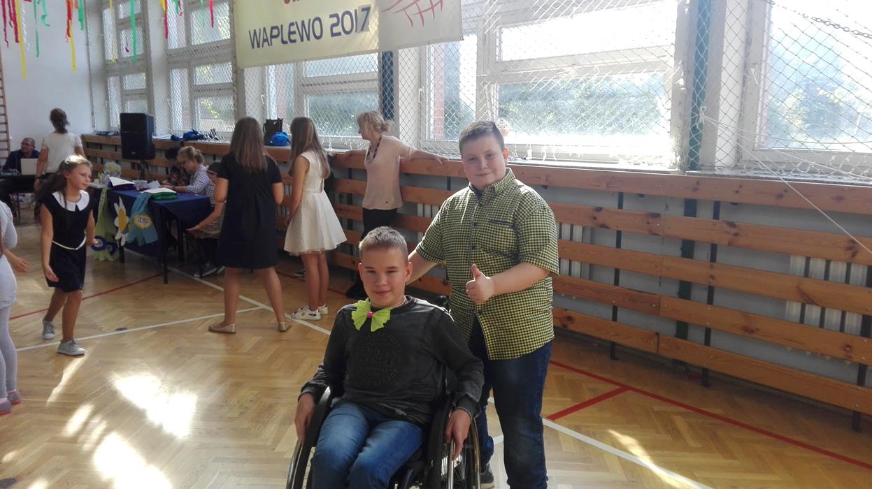 Zdjęcie uczniów w sali gimnastycznej podczas dyskoteki. Na przedzie dwóch chłopców, jeden poruszający się na wózku inwalidzkim