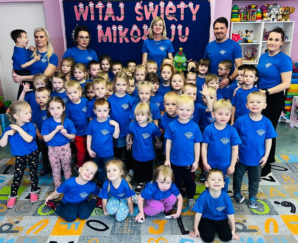 Liczna grupa dzieci uczęszczających do przedszkola, ich trzy wychowawczynie, pomoc nauczyciela i dyrektor stoją i pozują do zdjęcia pamiatkowego w niebieskich koszulkach z logo Przedszkola w Waplewie.