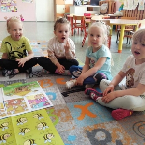 fot. A. Waśk. Chrońmy pszczoły.  Na zdjęciu grupa dzieci 3 – 4 letnich, siedzących na dywanie dookoła rozłożonych ilustracji dotyczących  pszczół. Dzieci wykonują ćwiczenia logopedyczne.