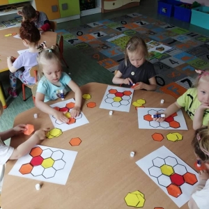 fot. A. Waśk. Chrońmy pszczoły. Grupa dzieci 3 – 4 letnich siedząca przy dwóch stolikach. Dzieci wykonują zakodowane plastry miodu, układają odpowiednio kawałki pomarańczowego, żółtego i czerwonego plastra.