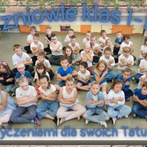 Liczna grupa uczniów klas 1-3 siedzi po turecku z sali szkolnej podczas śpiewania piosenki dla tatusiów.