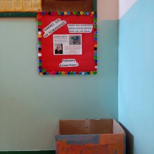 Plakat promujący akcję i karton z zebranymi nakrętkami umieszczone pod gazetką na korytarzu szkolnym.