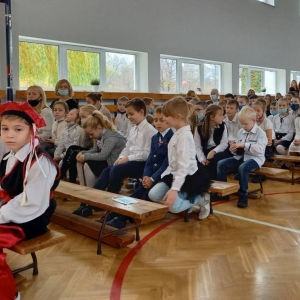 Zebrani - przedszkolaki, uczniowie i nauczyciele siedzą na ławkach i krzesełkach w sali gimnastycznej