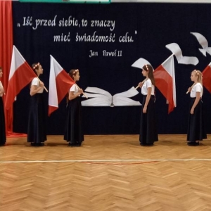 8 uczennic, w dwóch rzędach po 4 z każdej strony, stoi bogiem do dekoracji i bokiem do publiczności. Wszystkie ubrane są w białe koszulki, czarne długie spódnice, wianki z białych i czerwonych różek. Trzymają oparte na ramionach flagi Polski na drzewcach