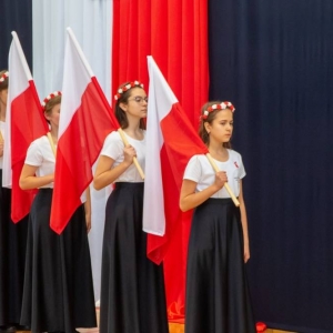 4 uczennice stoją bokiem do dekoracji i publiczności, trzymają oparte na ramionach flagi Polski na drzewcach