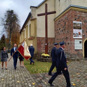 Przedstawiciele Rady Rodziców (2 kobiety i 1 mężczyzna) idą chodnikiem obok kościoła i niosą sztandar w asyście OSP w Waplewie