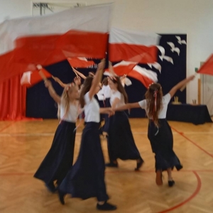 Taniec z flagami – 8 uczennic, w dwóch czwórkach kręci się w koło trzymając się za lewe dłonie, flagi trzymają uniesione nad głowami