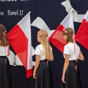 4 uczennice stoją bokiem do dekoracji i publiczności, trzymają oparte na ramionach flagi Polski na drzewcach