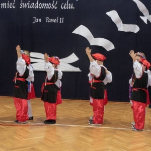 6 par przedszkolaków (dziewczynka z chłopcem) stoją ubrani w ludowe stroje, ustawieni w rzędzie gotowi do rozpoczęcia tańca -Krakowiaka. Chłopcy maja podniesione lewe ręce