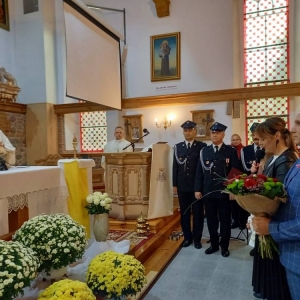 Przedstawiciele rodziców uczniów stoją przed ołtarzem, kobieta mówi do mikrofonu, mężczyzna trzyma wiązankę kwiatów