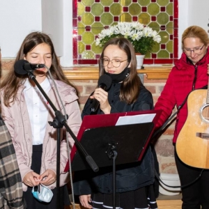 Trzy uczennice śpiewają w kościele do mikrofonu, za nimi kobieta z gitarą