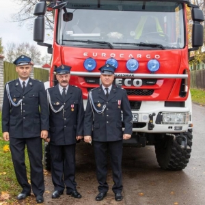 Trzech mężczyzn, asysta OSP w Waplewie w mundurach stoją przed wozem strażackim