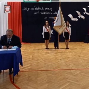 Burmistrz Olsztynka siedzi przy stoliku nakrytym niebieskim obrusem i podpisuje Akt Przekazania Sztandaru, za nim stoi poczet sztandarowy ze sztandarem