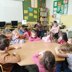 Uczniowie siedzą przy stolikach i słuchają czytanej przez nauczyciela książki.