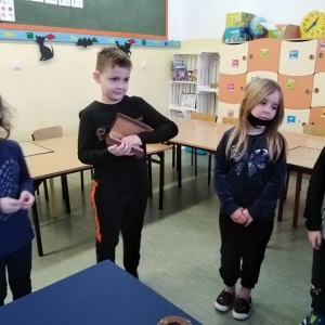 2 dziewczynki i 2 chłopców stoją w klasie szkolnej, jeden chłopiec trzyma drewnianą skrzyneczkę i losuje wróżbę