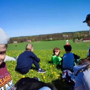 Grupa dzieci siedzi na trawie, w oddali stoi dziewczynka z aparatem w rękach i fotografuje otaczający ich krajobraz.