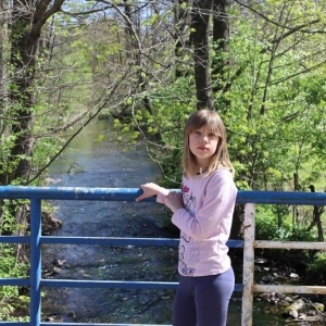 Dziewczynka pozuje do zdjęcia stojąc i opierając się o barierkę mostku nad rzeką.