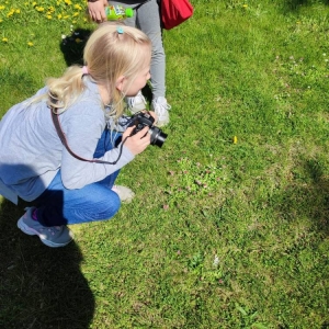 Na trawniku dziewczynka kuca z aparatem w rękach. Obok niej stoi jej koleżanka z klasy.