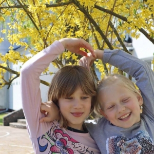 Dwie dziewczynki pozują do zdjęcia na tle kwitnącego na żółto drzewa. Uśmiechają się i łączą swoje dłonie nad własnymi głowami.