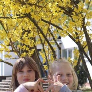 Dwie dziewczynki pozują do zdjęcia na tle kwitnącego na żółto drzewa. Swoje palce u dłoni układają w łączące się połówki serca.