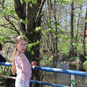 Chłopiec stoi tyłem do osoby fotografującej, opierając się na barierce mostku nad rzeką. Dziewczynka wrzuca do rzeki kamyk.
