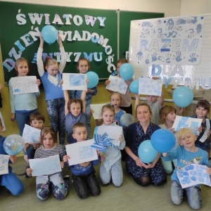 16 pierwszoklasistów i 2 nauczycielki ubrani na niebiesko, z balonami i pracami plastycznymi w barwach niebieskich, pozują do zdjęcia pamiatkowego.