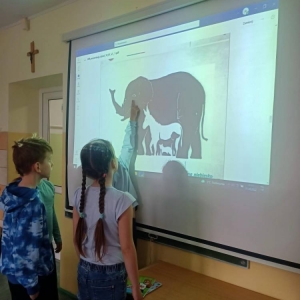2 uczennice i 2 uczniów oddziału II stoi przy wyświetlonej na projektorze prezentacji „Lekcja Autyzm” przygotowanej przez Fundację JiM – slajd z zadaniem „Jakie zwierzęta widzisz na obrazku?”. Jedna z uczennic palcem wskazuje na kontury słonia.