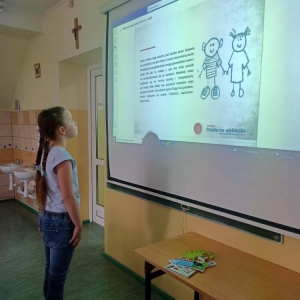 Uczennica oddziału II stoi przy wyświetlonej na projektorze prezentacji „Lekcja Autyzm” przygotowanej przez Fundację JiM – slajd z fragmentem książki dla dzieci pt. „Kosmita”.