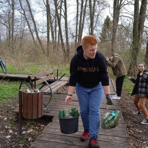 Ósmoklasiści sprzątają teren zielony w okolicy szkoły. Chłopiec idzie z wiaderkiem i koszykiem w rękach, pełnymi szklanych butelek
