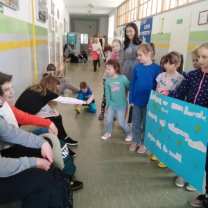 Grupa pierwszoklasistów i nauczycielka współorganizująca, z przygotowanym plakatem, podczas przerwy, na korytarzu szkolnym zachęca siódmoklasistów do przyłączenia się do akcji.