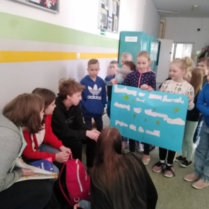 Grupa pierwszoklasistów, z przygotowanym plakatem, podczas przerwy, na korytarzu szkolnym zachęca uczniów klas 7 i 8 do przyłączenia się do akcji „Dzień bez zapalania światła”.