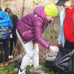 Szóstoklasiści sprzątają teren zielony w okolicy szkoły. Dziewczynka wrzuca śmieć do worka trzymanego przez jej koleżankę z klasy. Między ich nogami leżą znalezione butelki szklane.