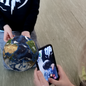 Chłopiec i dziewczynka siedzą na podłodze i poznają informacje o naszej planecie z wykorzystaniem smartfonu i interaktywnej karty edukacyjnej 4D.