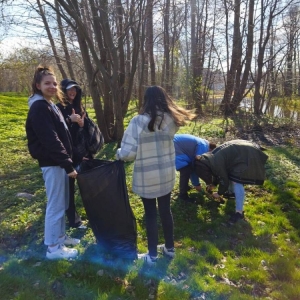 Siódmoklasiści sprzątają teren zielony nad rzeką w okolicy szkoły. Dwie dziewczynki trzymają duży czarny worek na śmieci.