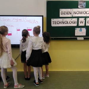 6 dziewczynek stoi przy tablicy interaktywnej. Zapisują daty i nazwy świąt majowych oraz rysują flagi Polski. Obok na tablicy kredowej, wywieszony wydrukowany napis: „Dzień Nowoczesnych Technologii w Edukacji”.