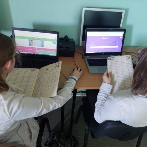 Podczas j. angielskiego, 2 uczennice siedzą przed laptopami, z książkami w rękach, na otwartej stronie z odmianą czasowników nieregularnych i rozwiązują quizy online.