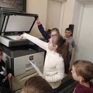 W sekretariacie, pierwszoklasistka wyciąga z drukarki biurowej, przygotowany do kserowania arkusz A4. Kilkoro dzieci stoi obok i się przygląda.