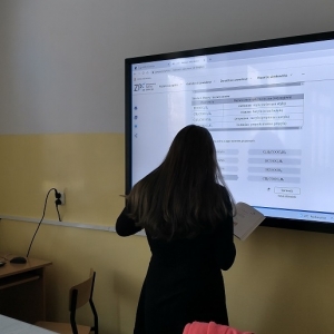 Ósmoklasistka, z książką w rękach, stoi i na telewizorze interaktywnym rozwiązuje tematyczne zadanie online udostępnione na Zintegrowanej Platformie Edukacyjnej.