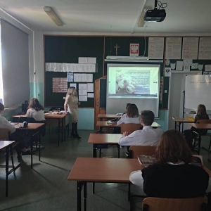 Siódmoklasiści siedzą pojedynczo w ławkach, nauczycielka j. polskiego stoi i prowadzi zajęcia. Na projektorze wyświetlona tematyczna interaktywna prezentacja Genially.