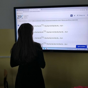 Ósmoklasistka stoi i na telewizorze interaktywnym rozwiązuje tematyczne zadanie online udostępnione na Zintegrowanej Platformie Edukacyjnej.