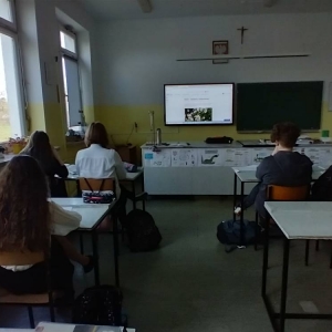 Ósmoklasiści, podczas zajęć chemii, siedzą w ławkach i oglądają wyświetlone na telewizorze interaktywnym tematyczne materiały edukacyjne online.