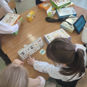 Rozłożone na stoliku pomoce dydaktyczne i tablet służące do wspomagającej metody komunikacji „Mówik”. Dwie dziewczynki na kalendarzu z symbolami pokazują symbol „wesoły”.