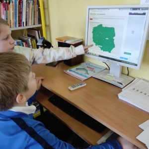 2 dzieci pracuje z wykorzystaniem komputera. Chłopiec siedzi, a dziewczynka stoi i palcem wskazuje na wyświetloną mapę Polski.