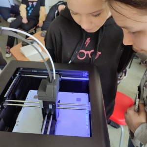 Dwie ósmoklasistki stoją, z pochylonymi nad drukarką 3D głowami, przyglądają się jak ona pracuje podczas drukowania.