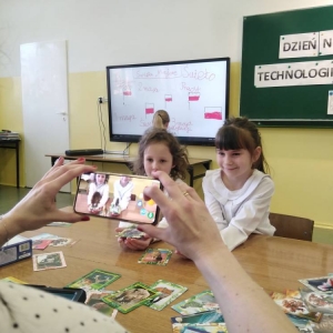 Dwie dziewczynki siedzą przy stoliku z kartami 4D na dłoniach i pozują do zdjęcia, które z wykorzystaniem aplikacji, robi im nauczycielka.