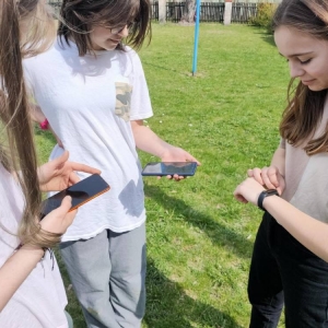 2 ósmoklasistki stoją na boisku szkolnym, ze smartfonami w dłoniach. Obok stoi ich koleżanka, ze smartwatchem na nadgarstku. Wszystkie patrzą na ekrany swoich urządzeń. 