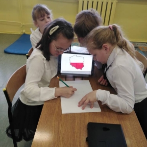 4 dziewczynki siedzą przy stoliku. Jedna z nich rysuje granice Polski w barwach biało-czerwonych, z wykorzystaniem projektora i tabletu.