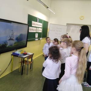 Grupa dzieci i nauczycielka stoją przed telewizorem interaktywnym z wyświetlonym filmem animowanym 3d. Uczennica i nauczycielka maja włożone okulary.