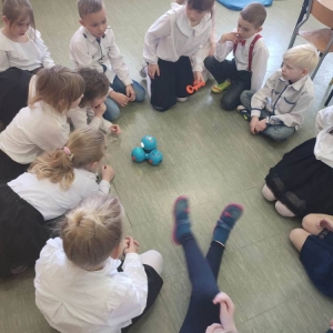Dzieci siedzą na podłodze po okręgu. Na środku porusza się robot Dash.